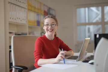 Jeune femme souriante assise à un bureau devant un ordinateur portable et tenant un crayon à la verticale sur une feuille