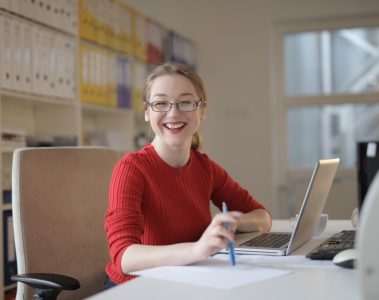 Jeune femme souriante assise à un bureau devant un ordinateur portable et tenant un crayon à la verticale sur une feuille
